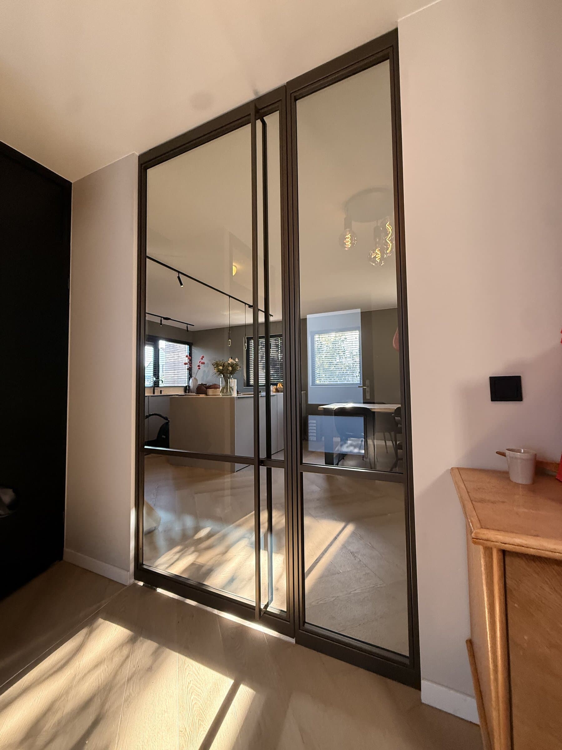 Kamerhoge stalen taatsdeur met vast zijlicht in anodic brown, gesloten, met helder glas en één dwarsregel, gezien vanuit de hal naar de woonkamer en keuken.