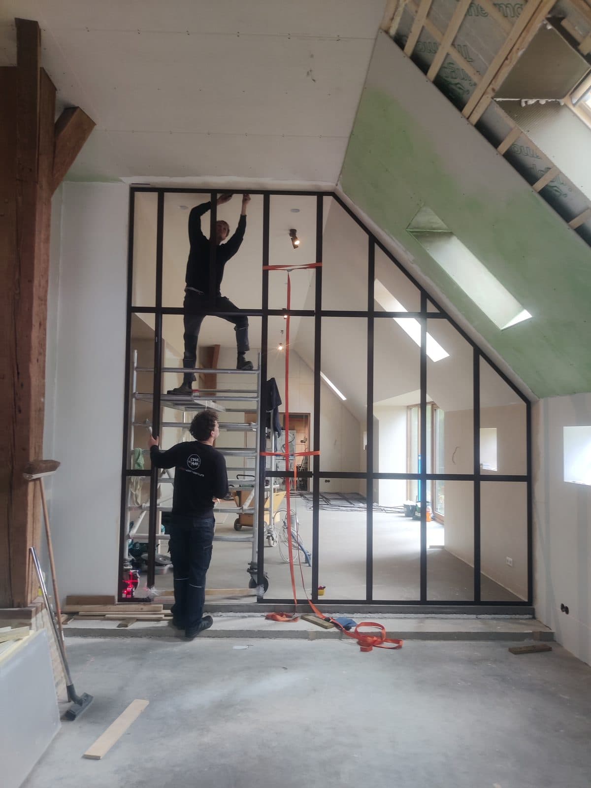 Twee monteurs van de StaalZaak plaatsen een kamerhoge stalen pui die met de schuine kap meeloopt, in een woning die nog in aanbouw is.