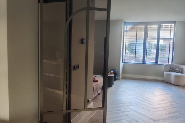 Kamerhoge bruine stalen scharnierdeur met een subtiel boogdesign en brons getint glas in een interieur met visgraatvloer.