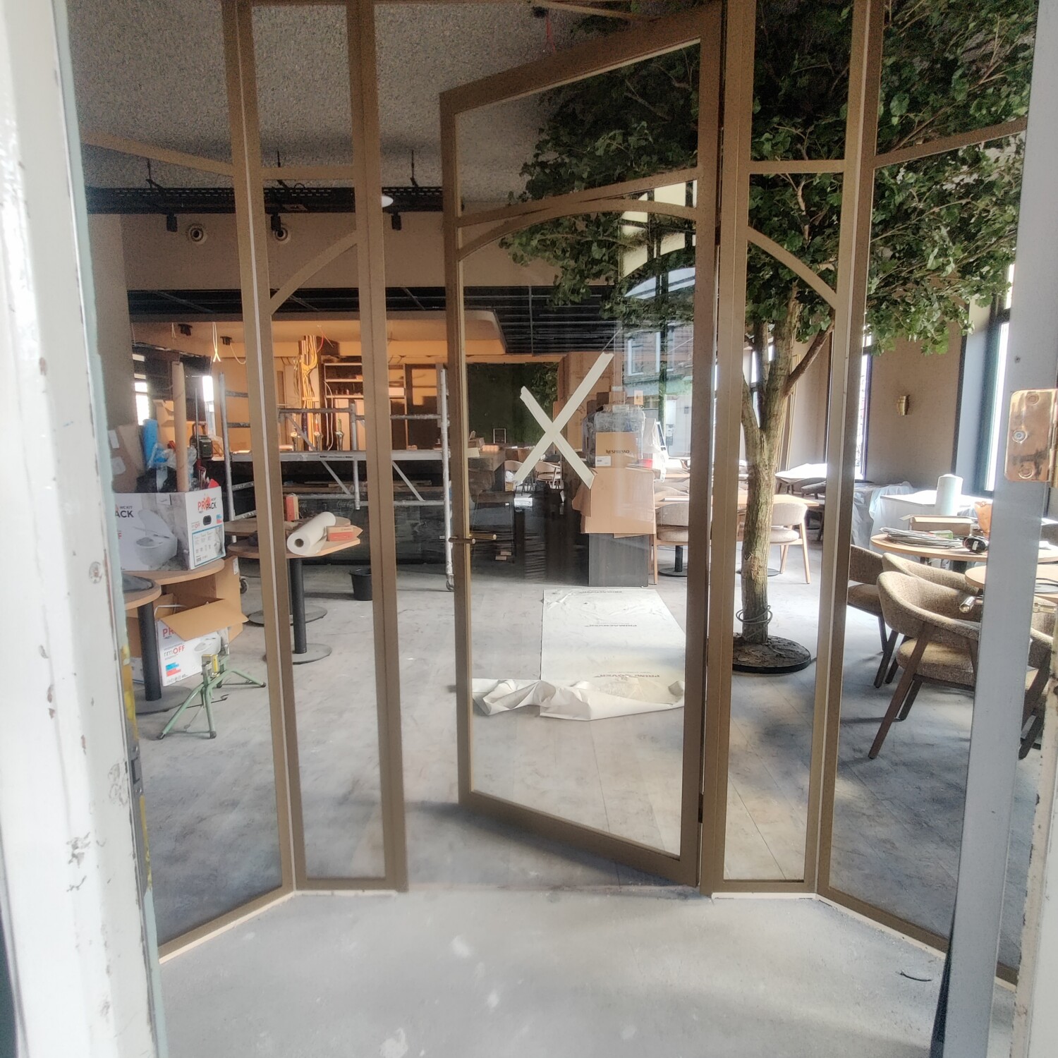 Op maat gemaakte goudkleurige stalen tochtsluis met een geopende binnendeur en helder glas in het interieur van een sfeervol café.