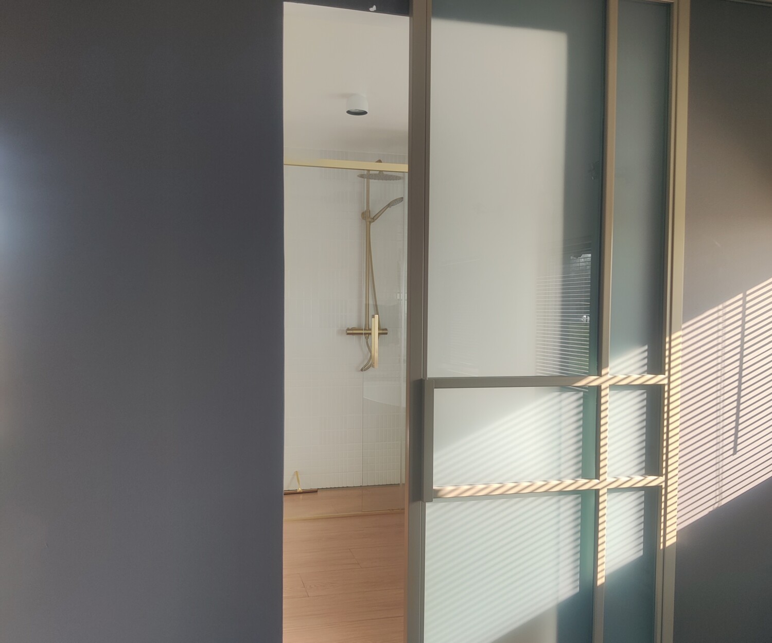 Geopende goud-bronskleurige stalen schuifdeur met frosted glas, met zicht op een moderne badkamer met gouden doucheset.