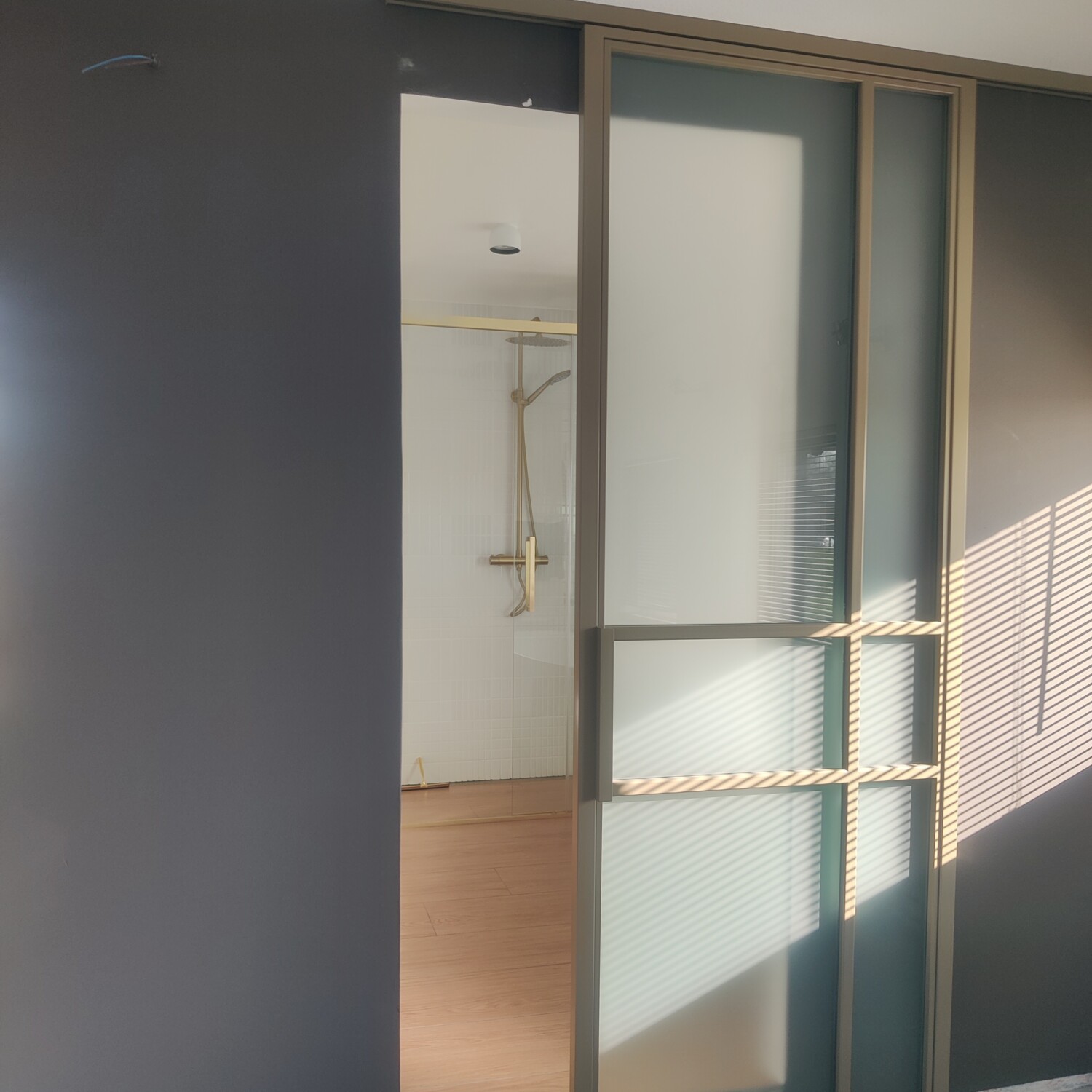 Geopende goud-bronskleurige stalen schuifdeur met frosted glas, met zicht op een moderne badkamer met gouden doucheset.