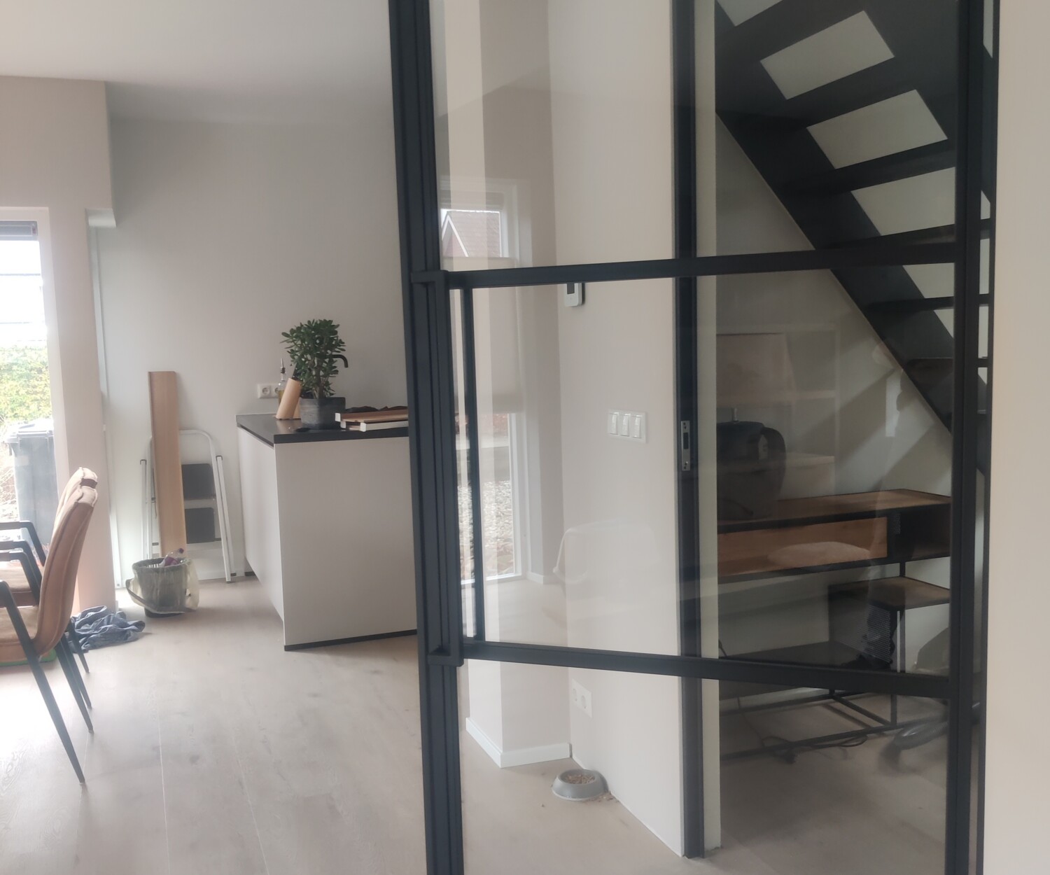 Lichte woonkamer met een openstaande zwarte stalen deur met helder veiligheidsglas en een strakke vlakverdeling.