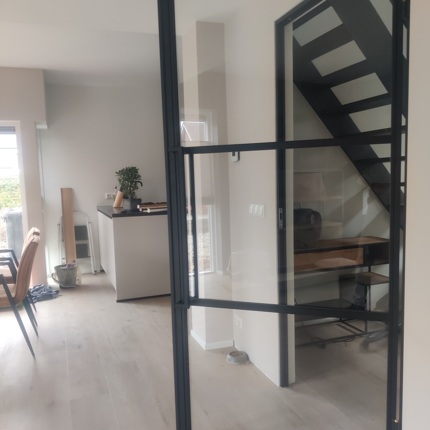 Lichte woonkamer met een openstaande zwarte stalen deur met helder veiligheidsglas en een strakke vlakverdeling.
