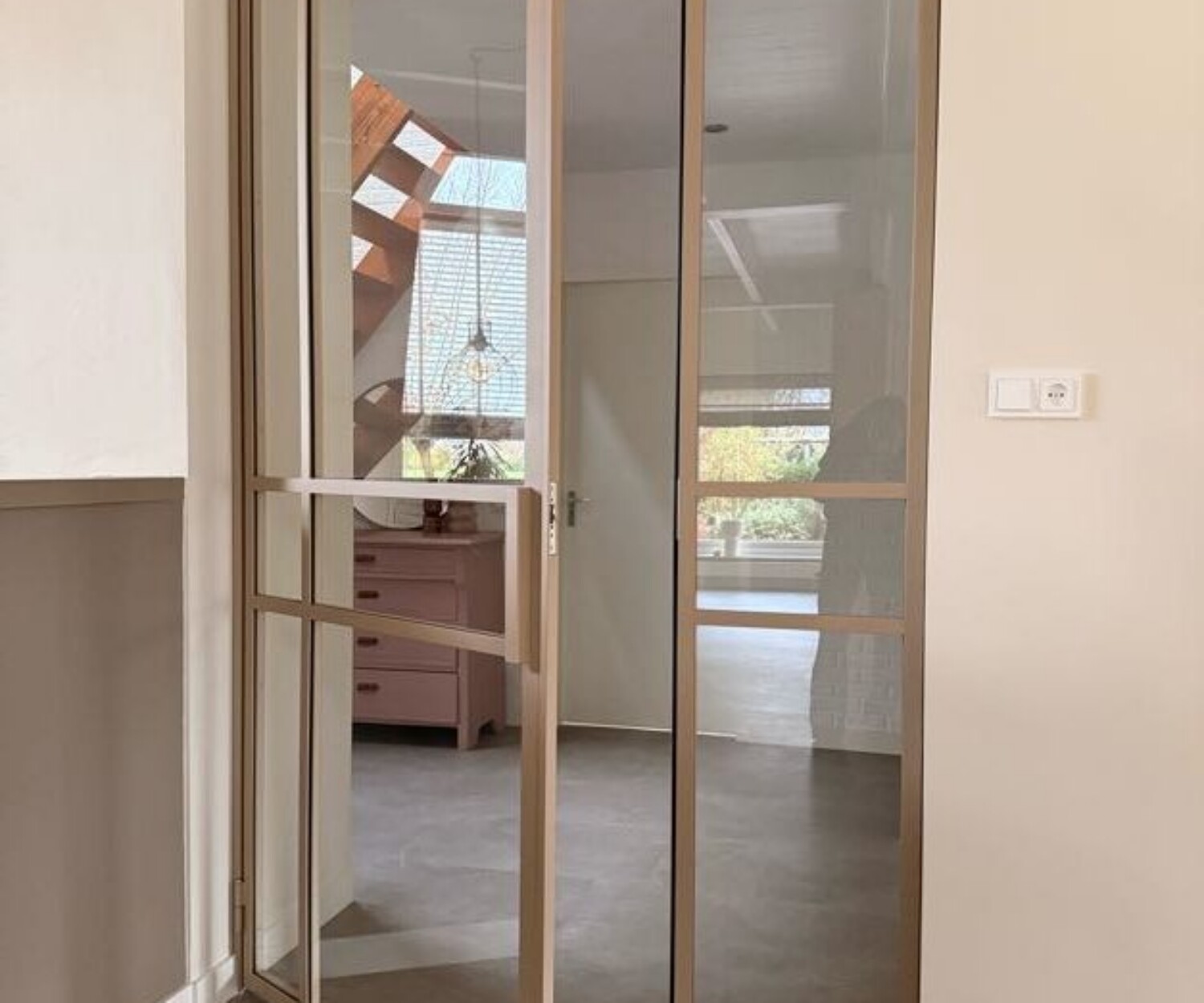 Kamerhoge beige stalen pui met een openstaande scharnierdeur, vast zijpaneel en helder glas gemonteerd in een licht interieur.