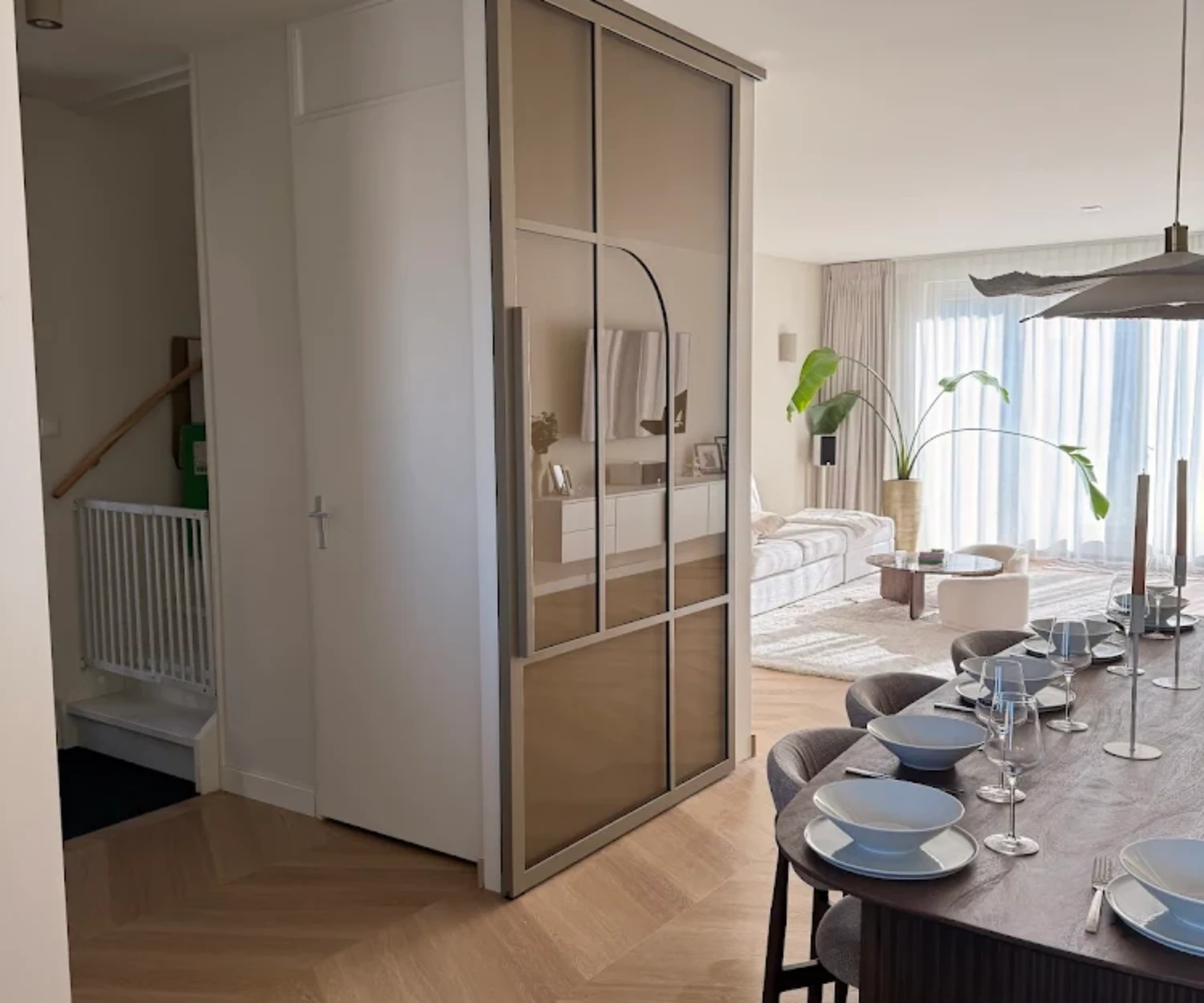 Luxe openstaande bronzen stalen binnendeur op rails in een sfeervolle woonkamer met visgraatvloer en houten eettafel.