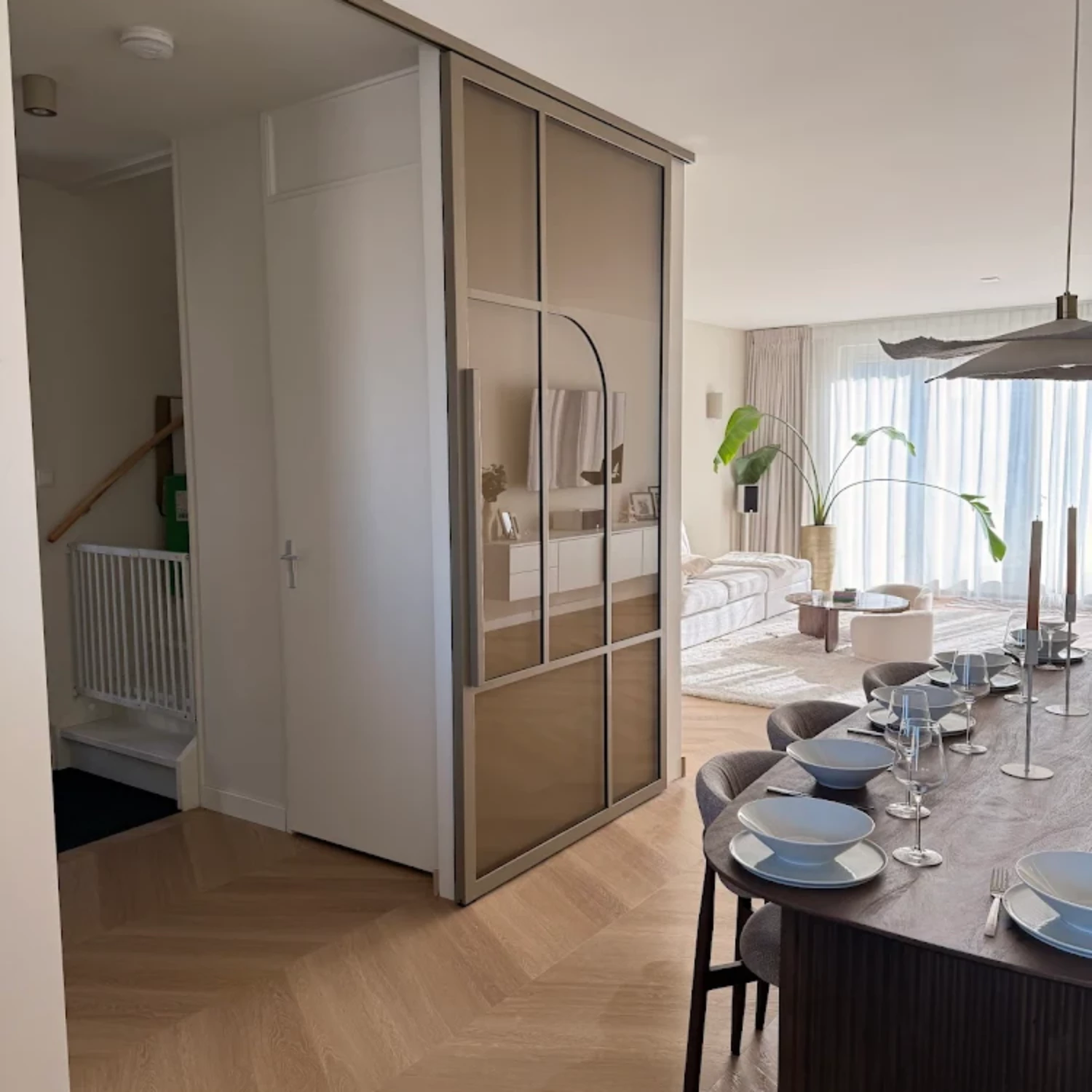 Luxe openstaande bronzen stalen binnendeur op rails in een sfeervolle woonkamer met visgraatvloer en houten eettafel.